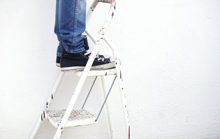 Person mit Jeans und schwarzen Turnschuhen steht auf einer Leiter und streicht